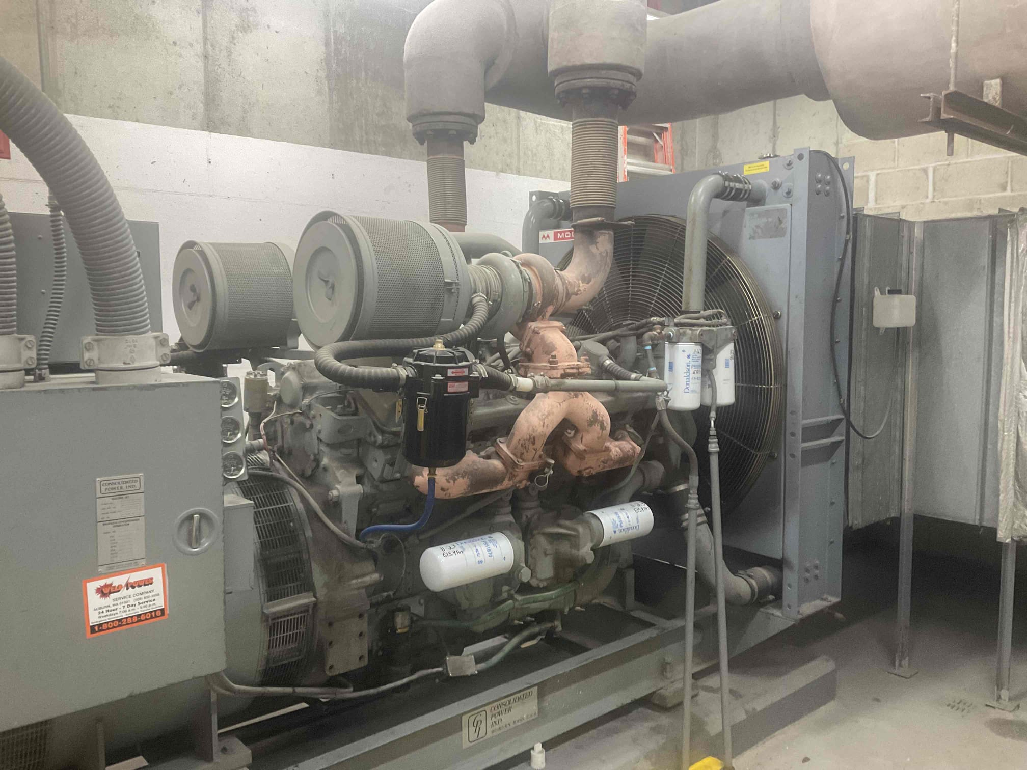 Commercial standby generator installed indoors with visible exhaust, electrical connections, and safety signage highlighting Generator hazards such as high voltage, hot surfaces, and moving mechanical components.