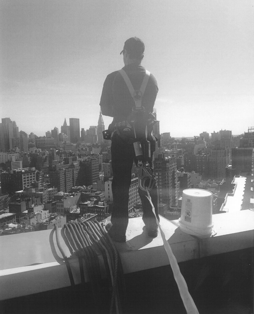  Generator technician on the roof of a building overlooking New York City with electrical cables for load bank testing and a bucket to collect waste oil when completing the preventative maintenance service on the rooftop generator. 